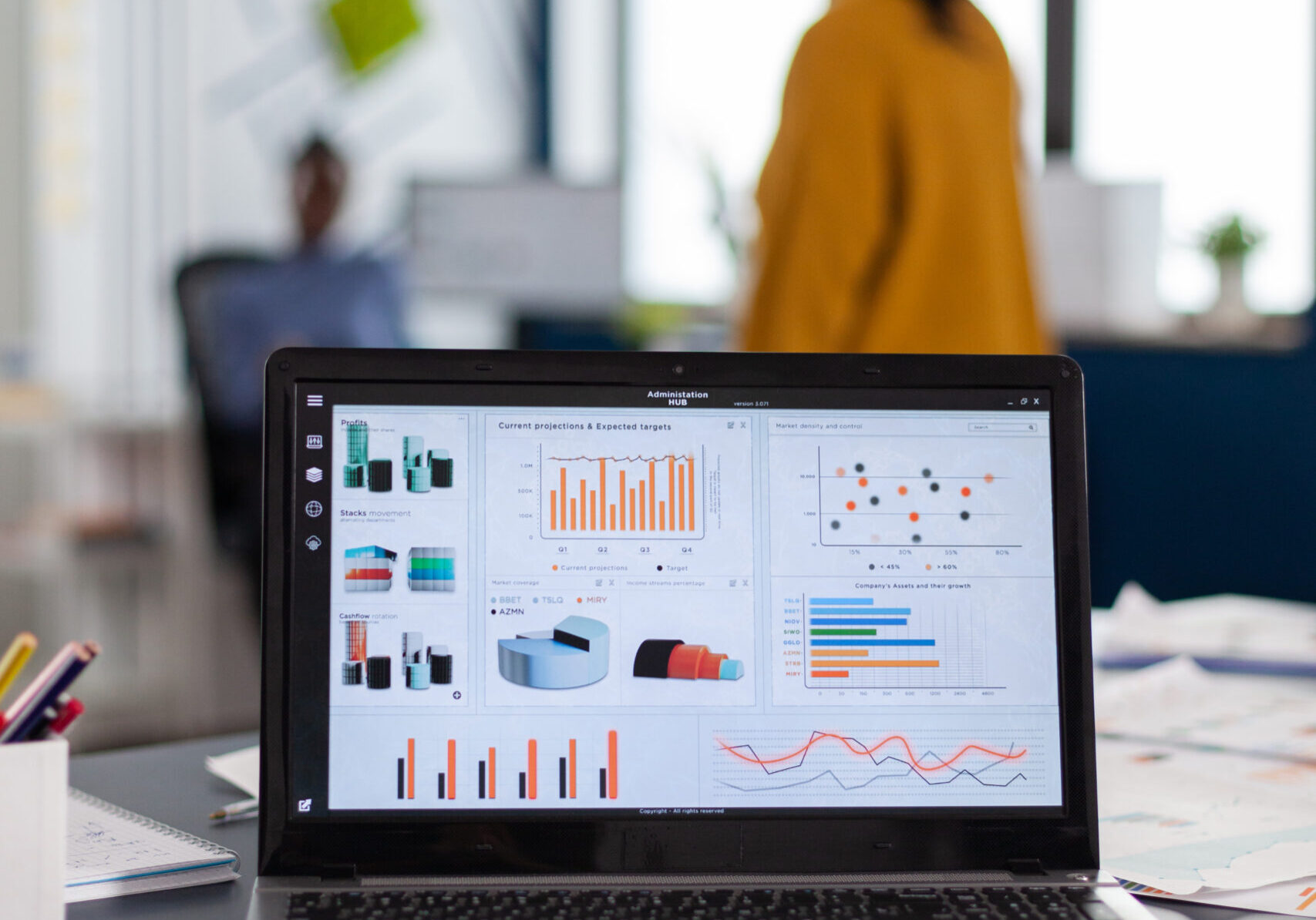 Close up of laptop with graphs, charts of marketing agency. Workplace area in business center with multi ethnic employees in it, shot of room with modern furniture and blue wall.
