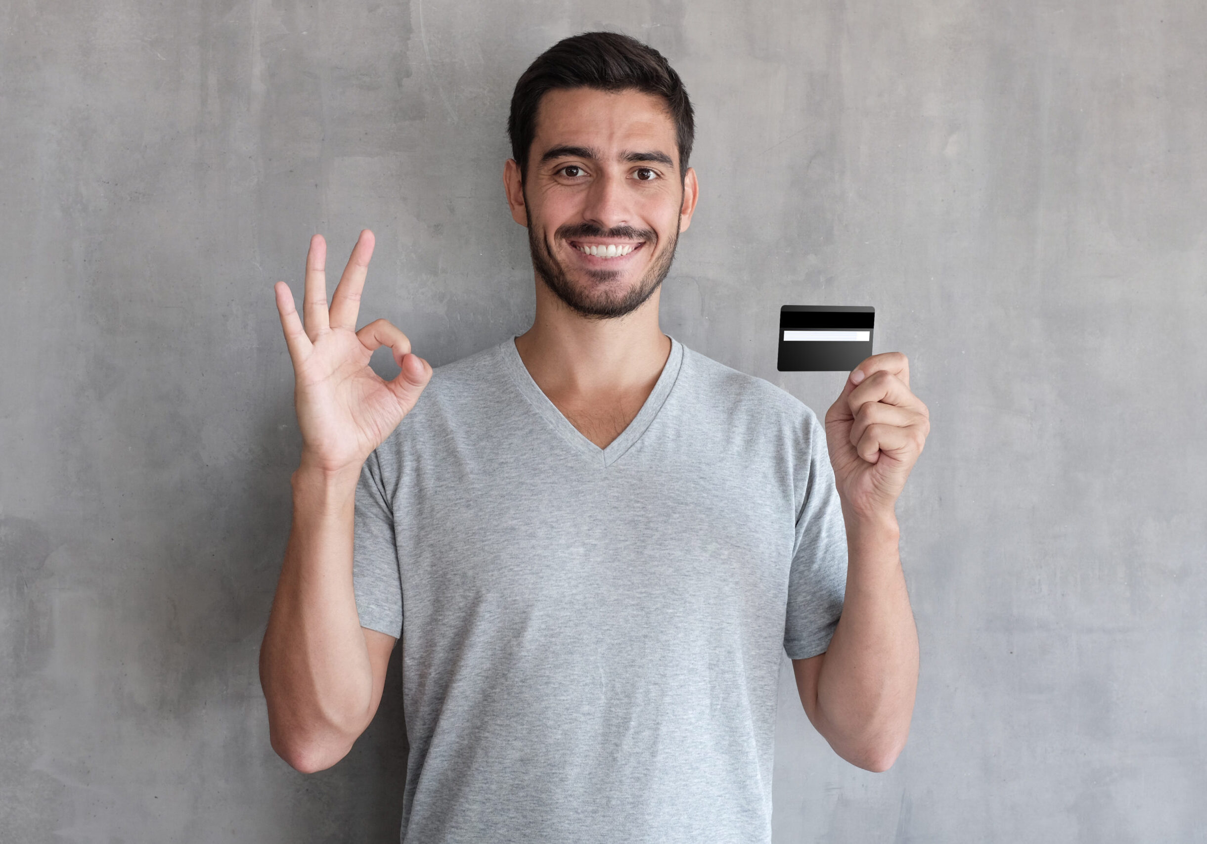 Portrait of young smiling man in t shirt, holding credit card and showing okay sign, standing against gray textured wall
